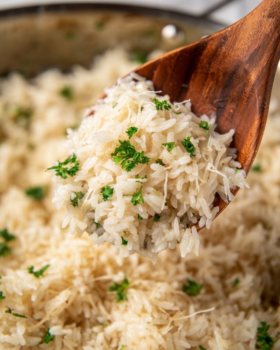 The image shows a close-up of cooked rice being lifted with a wooden spoon from a pan. The rice is white with a slight creamy texture, and it is garnished with small green parsley leaves scattered on top. There are thin shreds of melted cheese mixed into the rice, adding a soft and gooey layer. The background is blurred but shows more rice in the pan. The photo is framed tightly to focus on the spoonful of rice with the creamy, layered texture visible. photo taken with an iphone --ar 4:5 --v 7