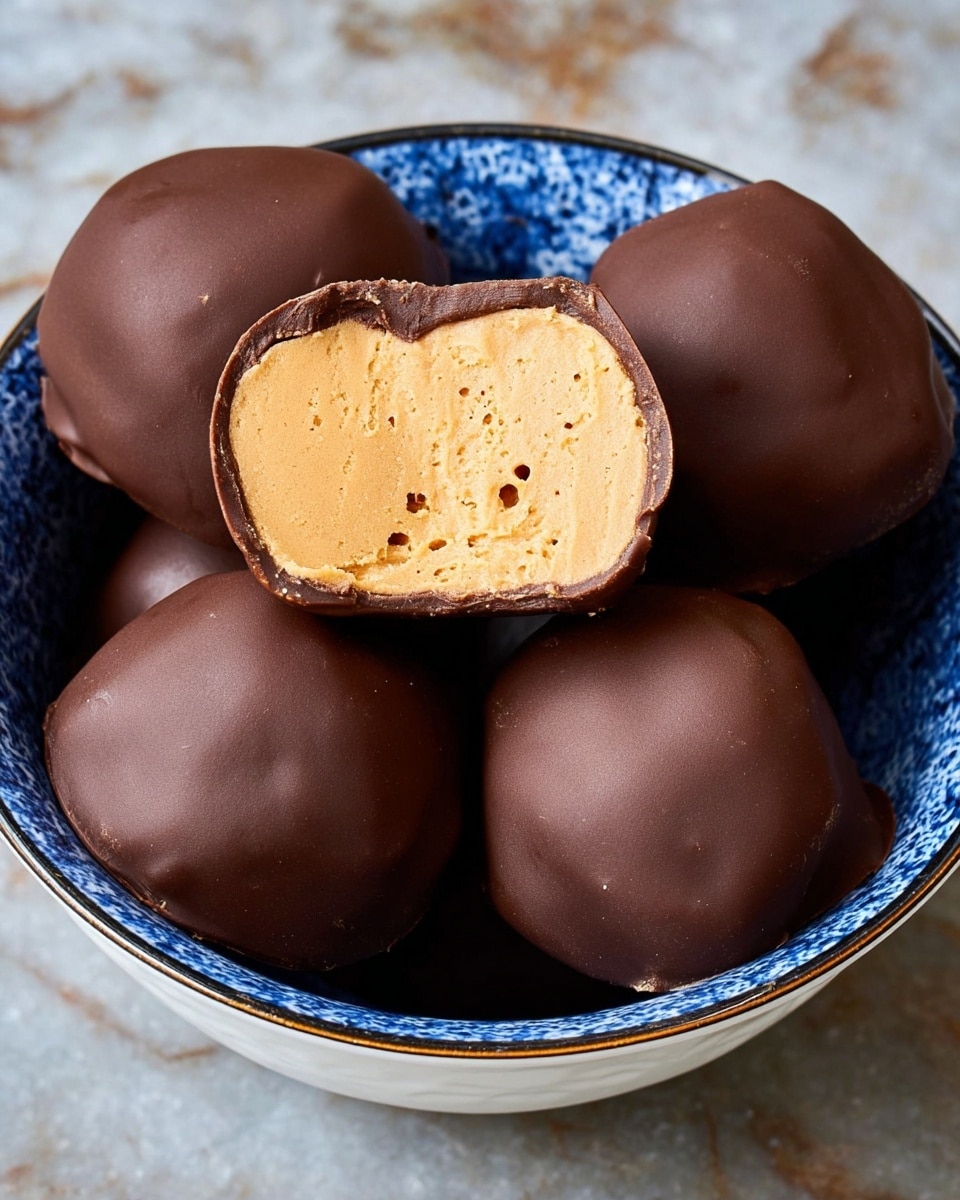 The image shows six round chocolate-coated peanut butter balls in a white bowl with blue patterns inside. Three of the balls are whole with a smooth, shiny, dark brown chocolate shell visible on top and around the sides, while the other three are cut in half, revealing a thick, creamy, light tan peanut butter layer with small air bubbles inside. The chocolate coating covers the peanut butter layer thinly but fully, creating a firm outer shell. The white bowl stands on a white marbled textured surface. photo taken with an iphone --ar 4:5 --v 7
