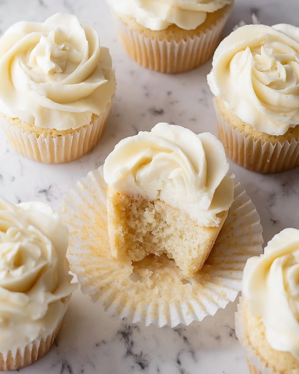 The image shows several vanilla cupcakes on a white marbled surface, each cupcake with one layer of light golden cake topped with one thick layer of creamy white frosting. The frosting is smoothly piped in a floral swirl pattern that creates a soft, petal-like texture. One cupcake in the center has its wrapper peeled back and is partially eaten, revealing the dense, moist texture of the cake inside. The cupcakes have white paper liners that loosely hold the cake. Photo taken with an iphone --ar 4:5 --v 7