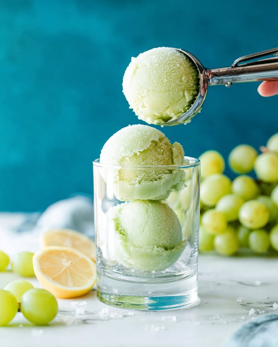 Two pale green scoops of sorbet with a soft, slightly grainy texture sit stacked inside a clear glass. A silver ice cream scoop held by a woman's hand places a third scoop above them. Around the glass on a white marbled surface are light green grapes and a wedge of lemon. In the background, a bunch of green grapes is softly blurred against a blue backdrop. Photo taken with an iphone --ar 4:5 --v 7