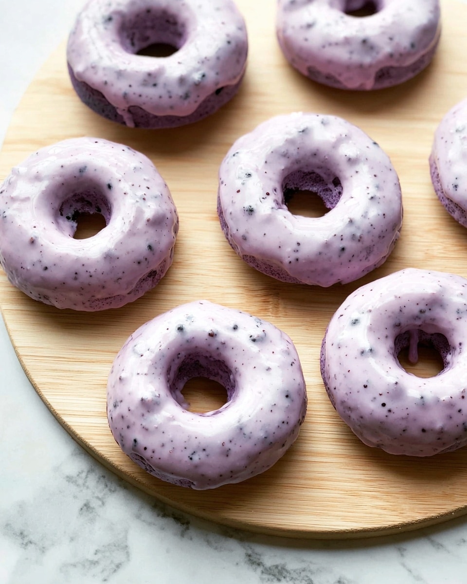The image shows six donuts placed on a light wooden board with a smooth texture. Each donut has one thick layer of pale purple glaze covering the top, with tiny darker purple spots spread across the glaze, giving it a slightly uneven surface. The donuts are arranged in a scattered pattern, without overlapping, and the wooden board sits on a white marbled surface. The donut's dough beneath the glaze is a darker color, hinting at possible berries or natural coloring in the dough itself. photo taken with an iphone --ar 4:5 --v 7