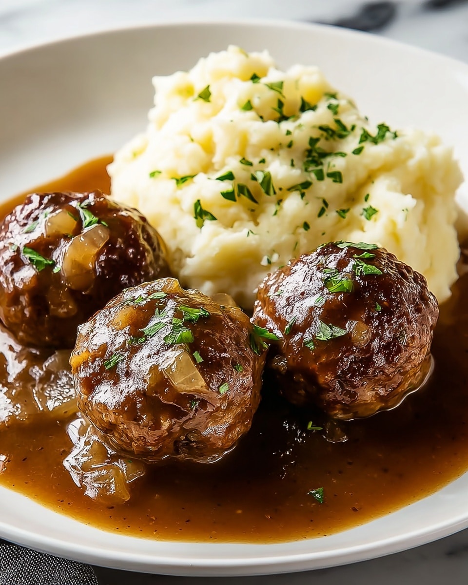 The image shows a white plate with three browned meatballs covered in a glossy brown gravy with visible chunks of onions, garnished with small green herbs on top. Behind the meatballs, there is a generous scoop of creamy mashed potatoes, also sprinkled with green herbs. The meatballs and potatoes rest on a pool of rich, thick brown gravy that slightly spreads around the edges of the plate. The plate sits on a white marbled surface. photo taken with an iphone --ar 4:5 --v 7