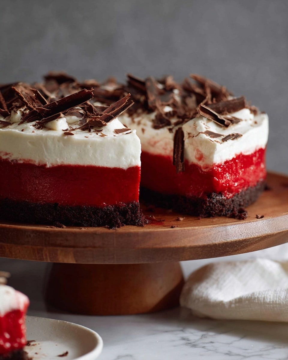 A three-layer dessert sits on a wooden stand against a gray background. The bottom layer is dark brown and crumbly like a cookie crust. The middle layer is smooth and bright red, thick and solid. On top, there is a thick layer of white whipped cream with a soft, fluffy texture. Thin, dark chocolate shavings are scattered over the whipped cream in an uneven pattern. In the lower left corner, part of the dessert is cut, showing the same layers inside. The surface under the stand is covered with a white marbled cloth. Photo taken with an iphone --ar 4:5 --v 7
