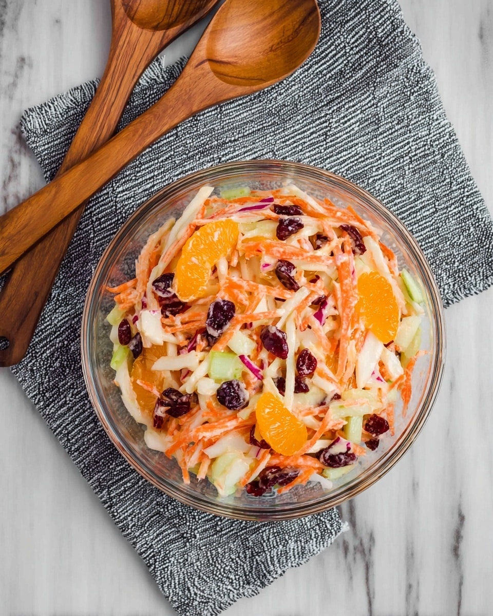 A clear glass bowl filled with a colorful salad sits on a white marbled surface. The salad has several layers: thin orange carrot strips, white apple slices, bright orange mandarin segments, green cucumber sticks, and dark red dried cranberries mixed evenly throughout. The salad looks fresh and slightly creamy with a light dressing coating all the ingredients. Next to the bowl, there are two wooden salad spoons resting on a neatly folded gray patterned cloth. photo taken with an iphone --ar 4:5 --v 7