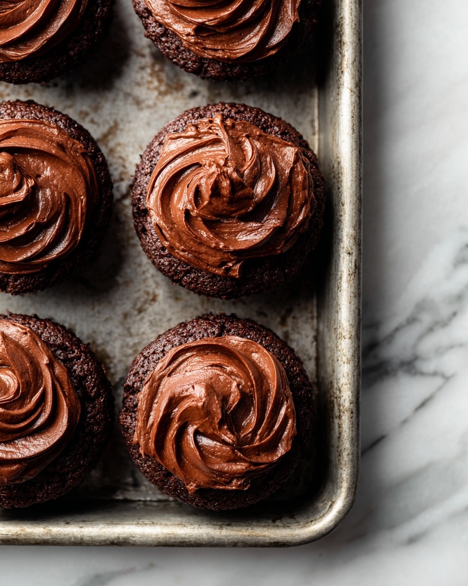 A close-up view of five chocolate cupcakes inside a metal baking tray, each topped with a thick layer of creamy, dark chocolate frosting swirled with soft, glossy texture. The cupcakes are dense and rich in appearance, with the frosting showing peaks and smooth curves that catch the light. The metal tray has a rustic look and is placed on a white marbled textured surface that adds a clean background contrast to the deep brown colors of the cupcakes and frosting. Photo taken with an iphone --ar 4:5 --v 7