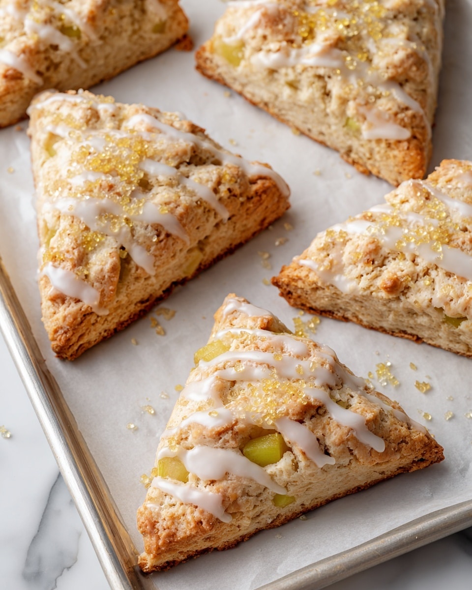 Several triangle-shaped scones are arranged on a baking tray covered with white parchment paper. Each scone has a light golden-brown color with visible chunks of pale green apple inside. The top layer is covered with a thin drizzle of white icing that looks smooth and slightly glossy. Golden sugar crystals are sprinkled over the icing, adding texture and sparkle. The tray is placed against a white marbled surface background. photo taken with an iphone --ar 4:5 --v 7