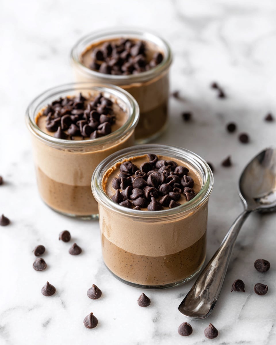 Three small clear glass jars filled with one layer of smooth light brown pudding topped with a thick layer of dark brown chocolate chips. The jars are placed closely together on a white marbled surface, with a silver spoon resting to the right side and scattered chocolate chips around them. The pudding's texture is creamy, and the chocolate chips are shiny and piled in the center of each jar. photo taken with an iphone --ar 4:5 --v 7