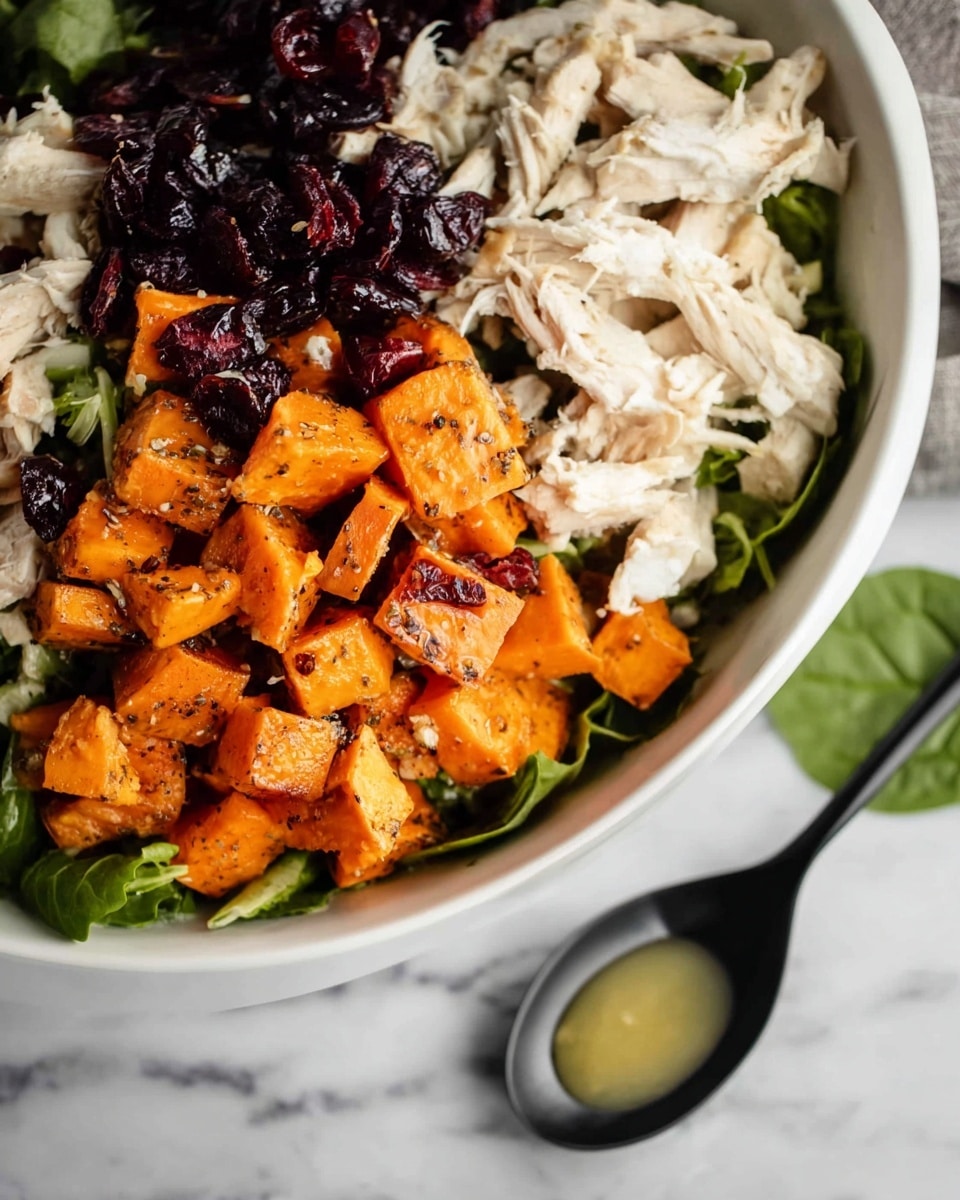 The image shows a white bowl filled with a layered dish, set on a white marbled surface. The bottom layer is fresh, green leafy lettuce. On top of this is a layer of bright orange roasted sweet potatoes, cut into small chunks with a slightly charred texture and black pepper seasoning. Next is a layer of dark purple dried cranberries, scattered over the sweet potatoes. The top layer consists of shredded white chicken breast pieces, which look tender and slightly seasoned. In the lower right corner, a black spoon with some light yellow sauce or dressing is lying on the surface next to a green spinach leaf. Photo taken with an iphone --ar 4:5 --v 7