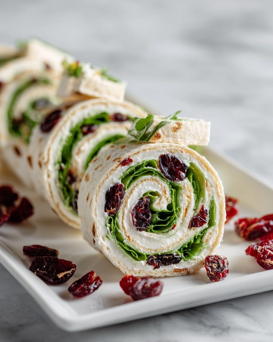 The image shows a stack of pinwheel sandwiches arranged on a white rectangular plate over a white marbled surface. Each pinwheel has three visible layers: the outermost layer is a soft tortilla, alternating between pale green and off-white colors; inside is a creamy white layer likely made from spreadable cheese; the innermost winding layer consists of fresh green lettuce leaves followed by scattered deep red dried cranberries throughout each roll. Some dried cranberries are also scattered around the plate on the white marbled surface, adding texture and color contrast. The image is bright and clearly focused on the detailed swirls of the pinwheels. photo taken with an iphone --ar 4:5 --v 7