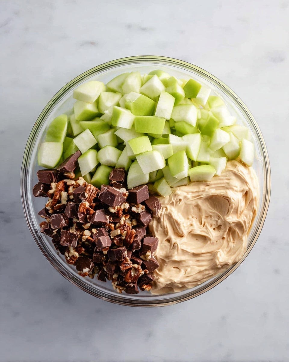 A clear round bowl is filled with three separate layers of ingredients arranged side by side: bright green chopped apples with a fresh, crisp texture on the top left; dark brown chocolate pieces with visible nuts inside on the bottom left; and a creamy, smooth, light beige layer of whipped mixture on the right side. The bowl is set on a white marbled surface, showing the contrast in colors and textures clearly. photo taken with an iphone --ar 4:5 --v 7