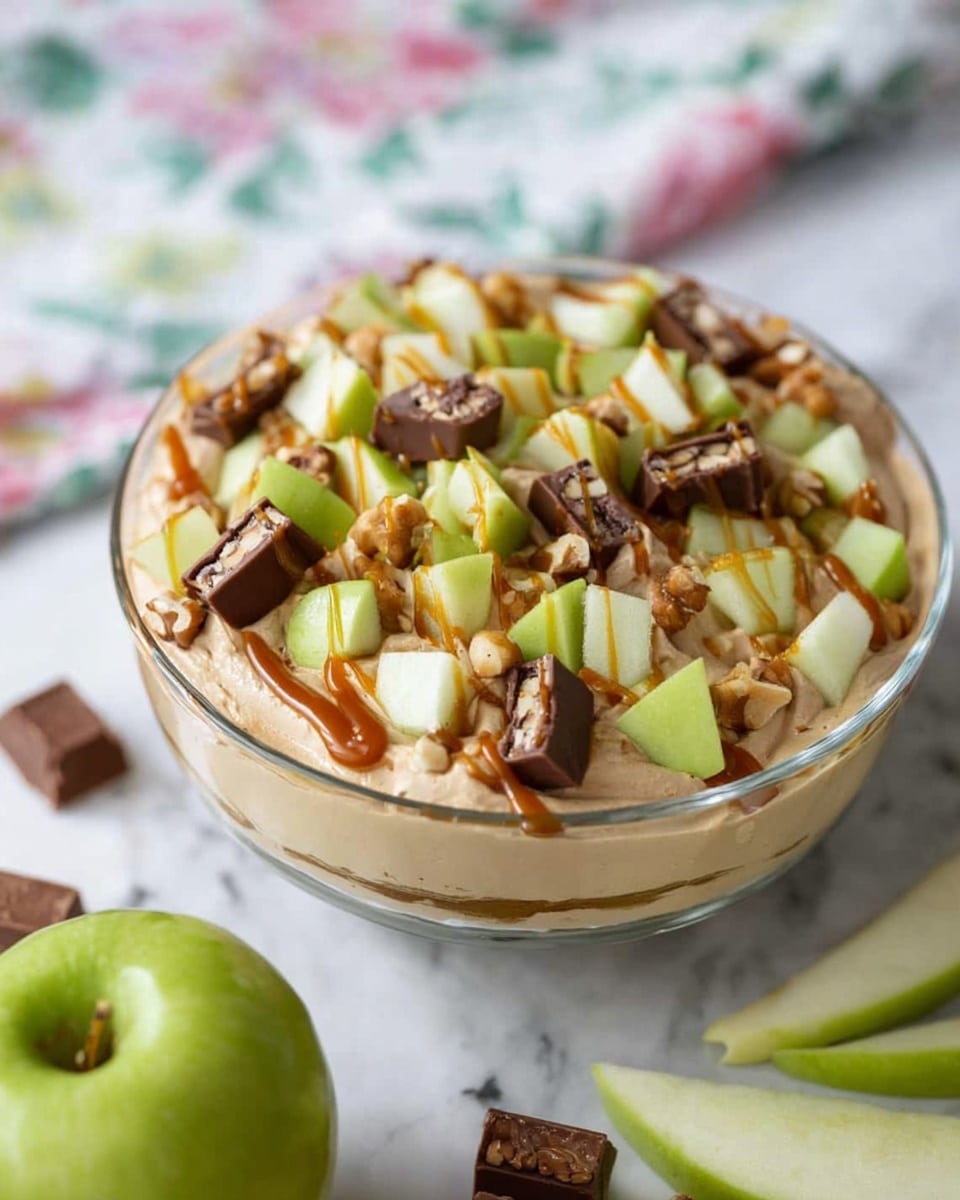 A clear glass bowl filled with a creamy, light brown mousse as the base layer, topped with chopped green apple pieces that are fresh and crisp-looking. Scattered over the apples are small chunks of chocolate and nut candy bars with visible nuts inside. A drizzle of caramel sauce is spread over the top, enhancing the colors and texture. The bowl is placed on a white marbled surface with a green apple and sliced candy bars near it, while a cloth with a floral pattern is blurred in the background. Photo taken with an iphone --ar 4:5 --v 7