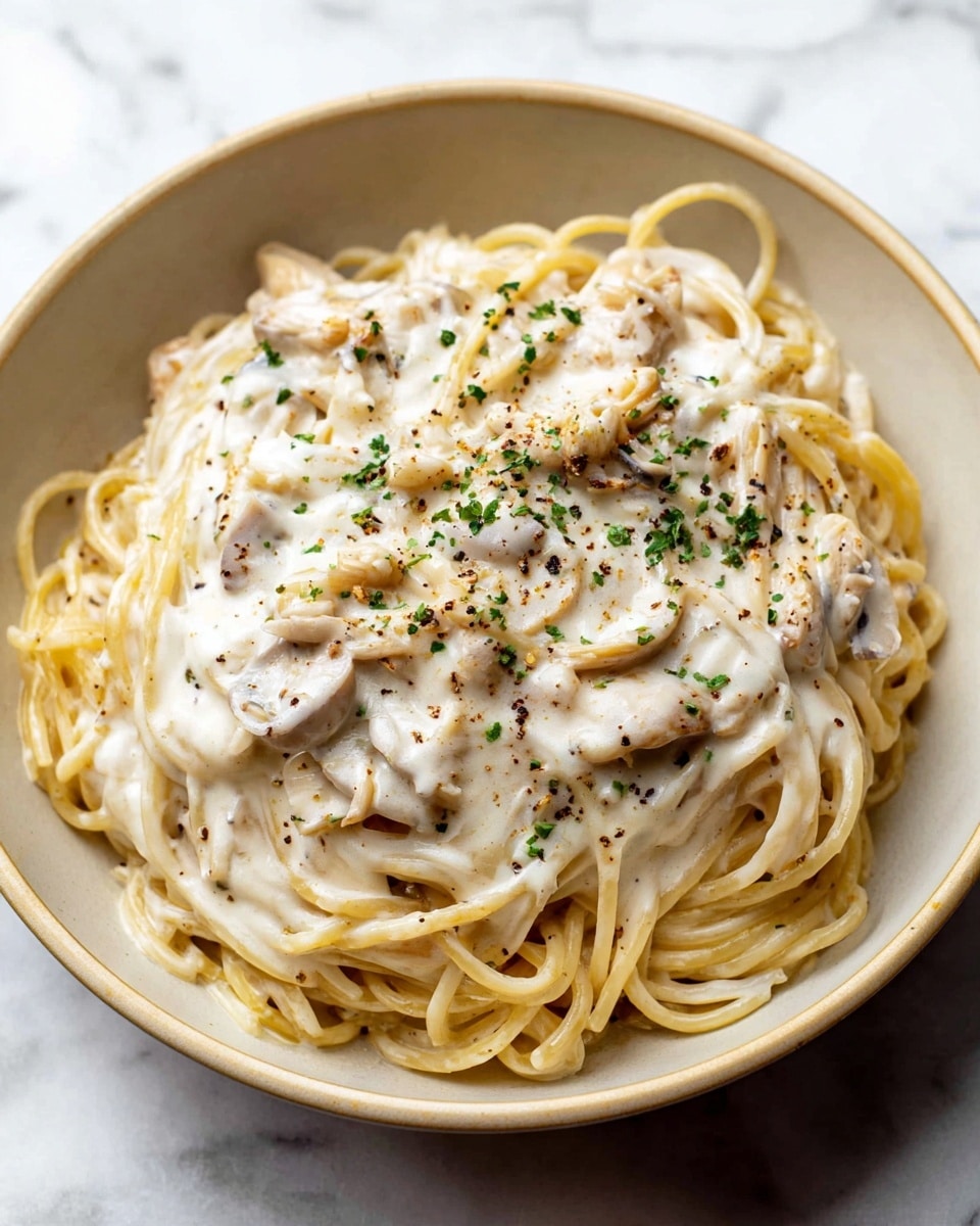 A close-up view of a bowl of creamy white pasta with spaghetti noodles covered in a thick, smooth white sauce mixed with small pieces of what looks like chicken or mushrooms. The sauce is sprinkled evenly with small green herbs and black pepper bits, adding specks of color on top. The bowl is white with a light beige interior, placed on a white marbled surface. The dish has one layer of pasta with sauce coating it thoroughly. photo taken with an iphone --ar 4:5 --v 7
