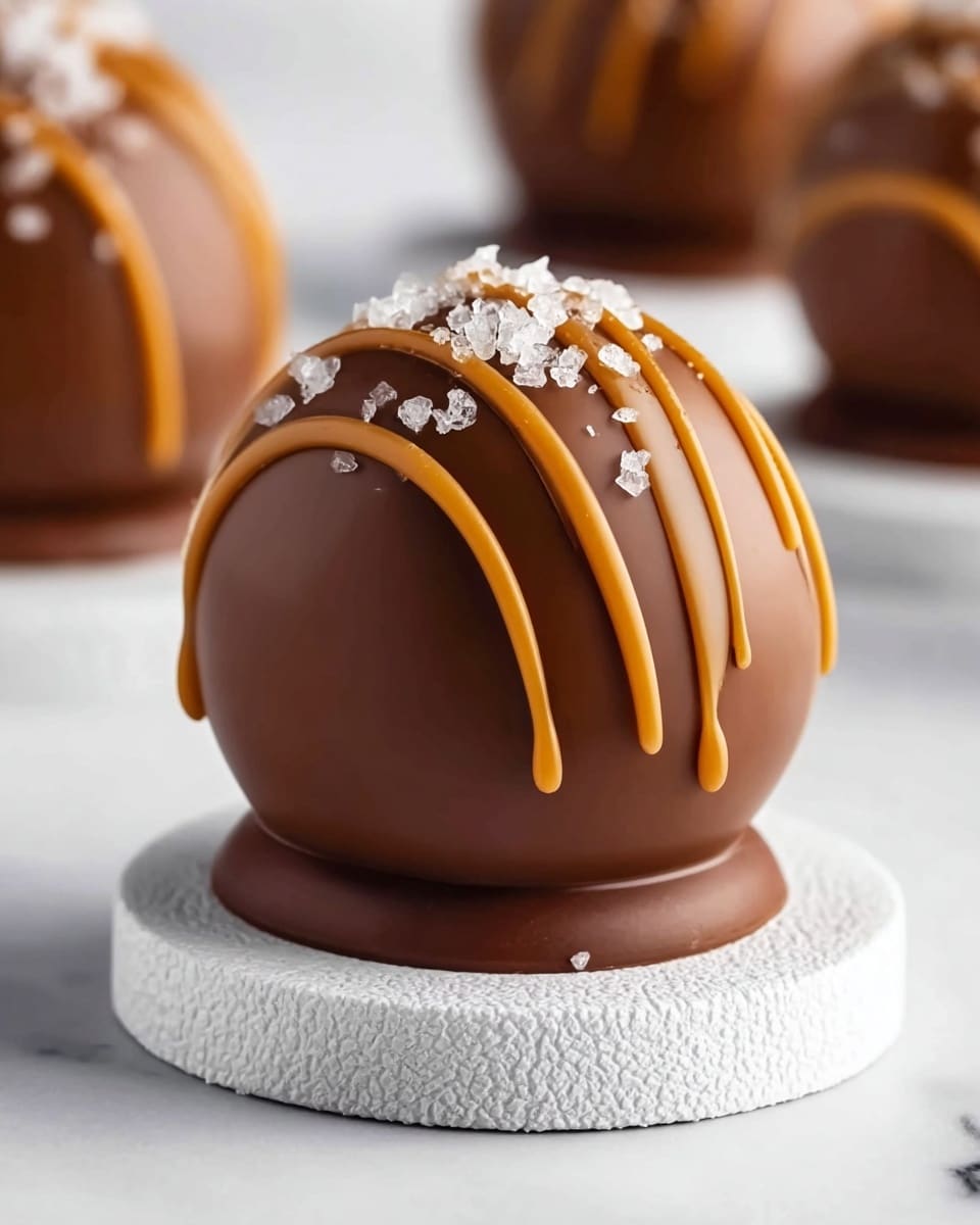 A smooth, round chocolate sphere sits on a small, flat chocolate base; the sphere is coated in a rich brown chocolate and decorated with three evenly spaced caramel-colored stripes running from top to bottom. On the top middle section, crushed white salt crystals are sprinkled, adding texture and contrast. The sphere rests on a white textured coaster that lies on a white marbled surface, with more chocolate spheres blurred softly in the background. photo taken with an iphone --ar 4:5 --v 7