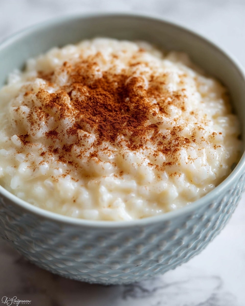 A close-up of a glass bowl filled with creamy white rice pudding, showing a thick, textured layer of soft rice grains in a smooth, milky base. The pudding is topped with a light sprinkle of brown cinnamon powder and a few thin, white coconut flakes on the surface. The bowl sits on a white marbled surface, and a gold spoon is positioned next to the bowl. photo taken with an iphone --ar 4:5 --v 7