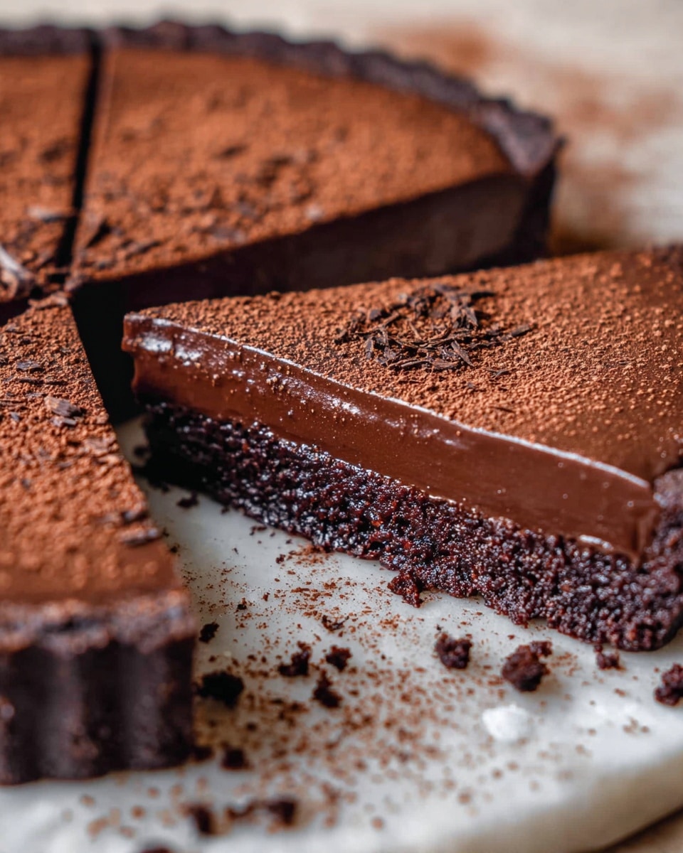 A round chocolate tart with a smooth and shiny dark brown top layer covers the entire tart, except for one slice that reveals a slightly softer chocolate layer beneath. The tart is dusted with cocoa powder on half of the surface, creating a textured light brown contrast against the glossy dark chocolate. Two bright red strawberries with green leaves sit on top of the tart, one whole and one sliced, placed on the chocolate side near the cut slice. The tart sits on crumpled parchment paper, with a white bowl filled with cocoa powder nearby and a metal sieve resting above the scene, all on a white marbled surface. Photo taken with an iphone --ar 4:5 --v 7