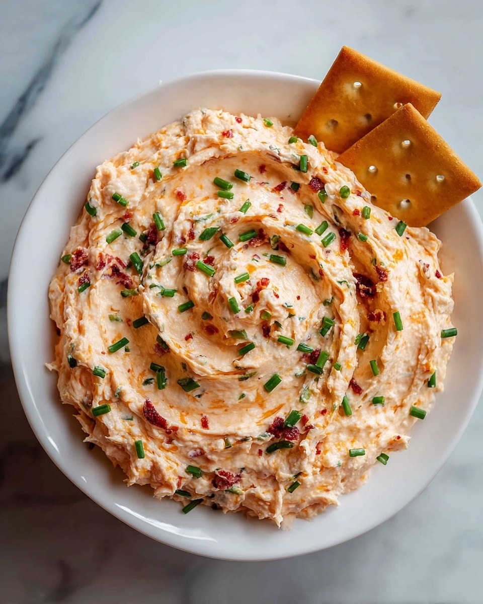 A white bowl is filled with a creamy spread that has a light orange color mixed with small red bits and green chives evenly spread throughout. The spread is swirled in a circular motion, creating a textured look with some peaks. Two square, golden-brown crackers rest at the edge of the bowl on a white marbled surface. photo taken with an iphone --ar 4:5 --v 7