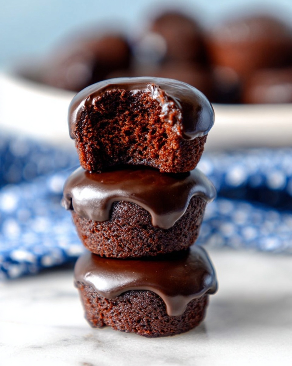The image shows three small chocolate cakes stacked on top of each other on a white marbled surface. Each cake is covered in thick, glossy dark chocolate glaze that slightly drips down the sides. The top cake has a bite taken out of it, revealing its moist, rich, dark brown inside. In the background, more chocolate-covered cakes and a blue and white cloth can be seen slightly blurred. photo taken with an iphone --ar 4:5 --v 7