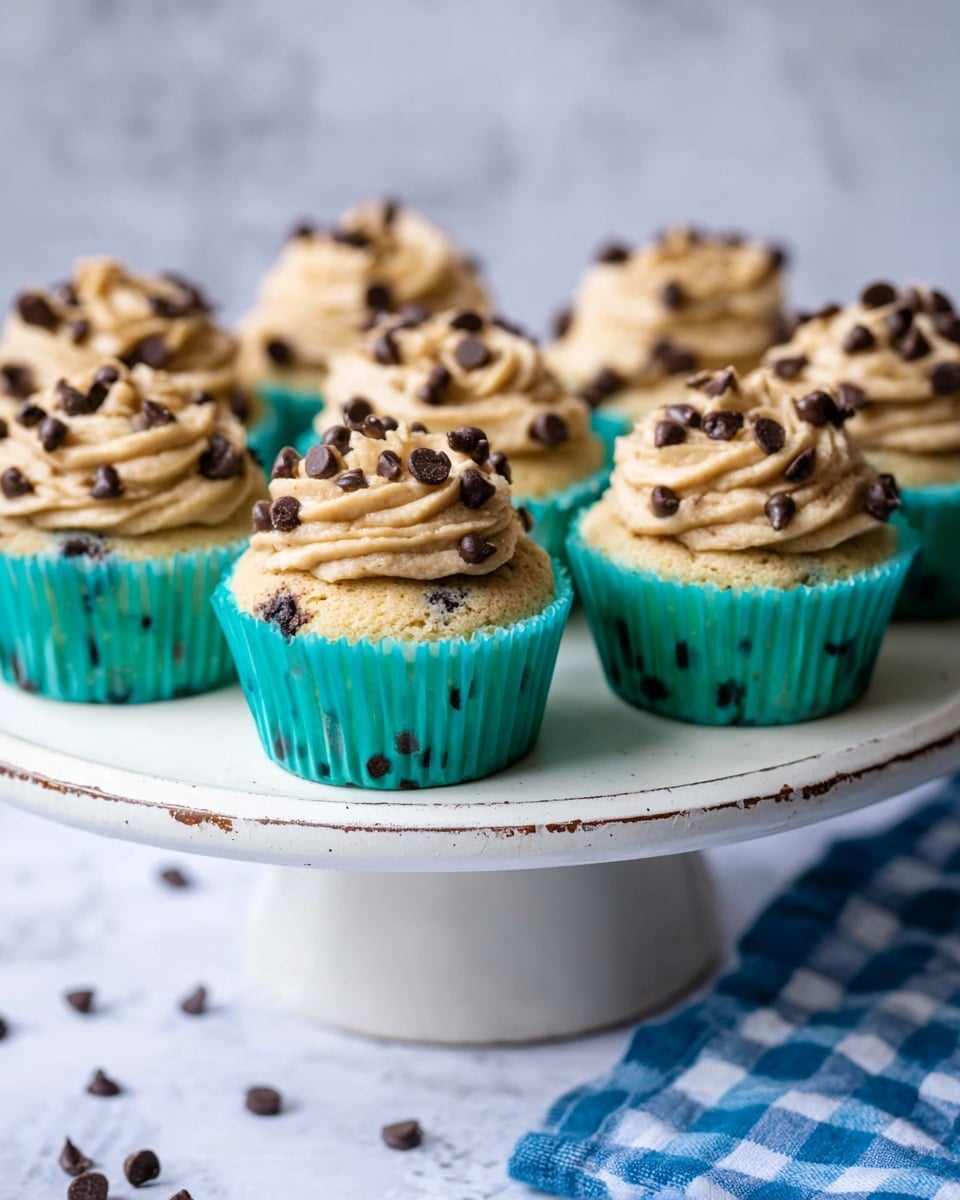 The image shows several cupcakes arranged on a white cake stand with a slightly chipped edge. Each cupcake is in a turquoise paper liner and topped with two layers of beige frosting that has a creamy, smooth texture, studded with small and large dark chocolate chips scattered all over the frosting and visible in the cake itself. The cupcakes are set against a white marbled texture surface with a blue and white checkered cloth partially visible at the bottom. Photo taken with an iphone --ar 4:5 --v 7