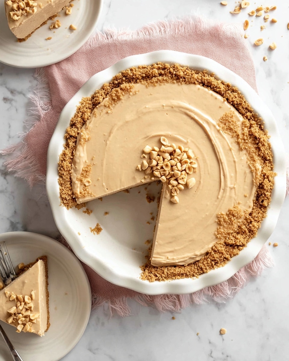 A creamy peanut butter pie sits in a white pie dish with scalloped edges on a white marbled surface. The pie has two main layers: a light tan, smooth and creamy peanut butter filling on top, and a crumbly golden-brown cookie crust lining the bottom and edges. The center of the pie is topped with a small pile of chopped peanuts. One slice has been cut and partially lifted, showing the filling and crust clearly. Nearby, a white plate with a slice of the pie and a silver fork is placed, with some scattered peanuts around it. There is a soft pink cloth with frayed edges under the pie dish. photo taken with an iphone --ar 4:5 --v 7