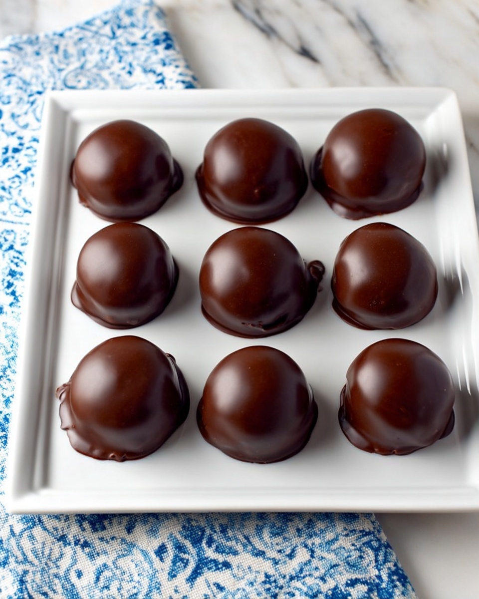 Nine round chocolate-covered treats are neatly arranged in three rows of three on a large white square plate. Each treat has a smooth, shiny dark brown chocolate coating that glistens under the light, with a soft, rounded dome shape. The treats sit closely but not touching, and the chocolate coating slightly drips over the edges of some, adding texture. The plate rests on a white marbled surface with a blue and white patterned cloth partially underneath, adding contrast to the simple, elegant arrangement. photo taken with an iphone --ar 4:5 --v 7