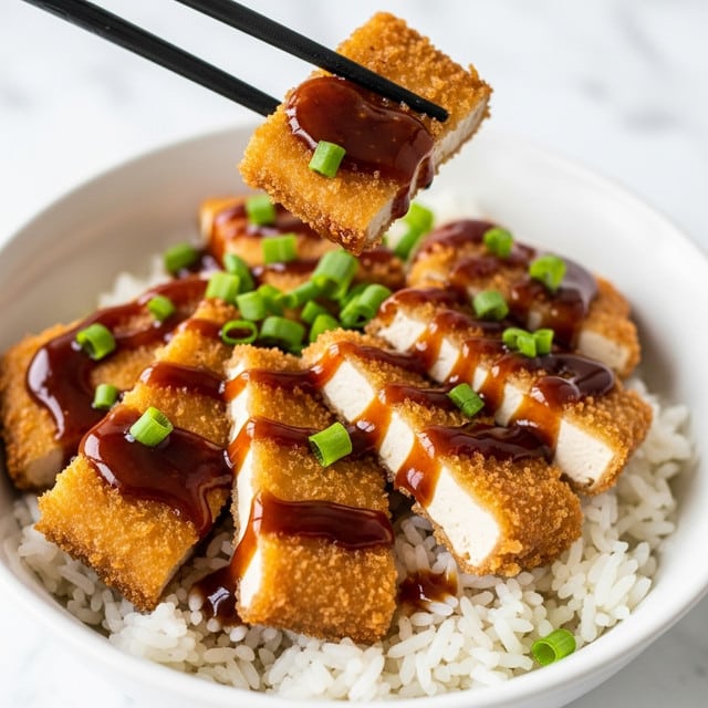 A white bowl contains a bed of white steamed rice as the bottom layer, topped with several pieces of golden-brown crispy fried chicken cut into thick slices showing the white inside. The chicken is covered with a shiny dark brown sauce that glistens under the light and is garnished with thinly sliced green onions and seaweed strips. A pair of black chopsticks, held by a woman's hand, is picking up one piece of chicken, highlighting the texture of the crispy crust and moist meat. The background and surface are white marbled texture. photo taken with an iphone --ar 4:5 --v 7