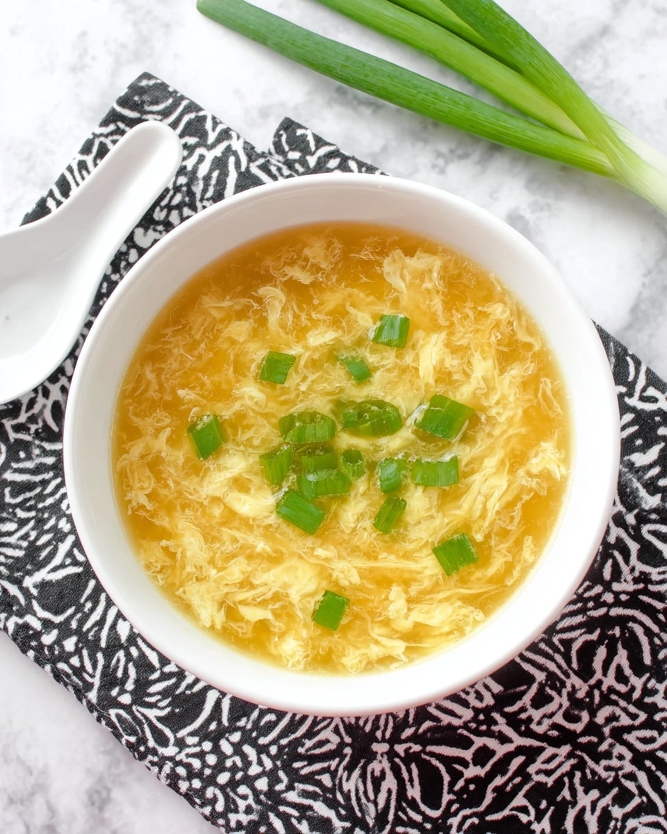 A white bowl filled with a light golden egg drop soup showing soft, wispy egg ribbons floating in a clear broth, topped with small bright green chopped scallions scattered centrally. The bowl is placed on a white marbled surface with a black geometric patterned cloth underneath. Nearby, a white ceramic spoon rests on the surface beside the bowl, and two fresh green scallion stalks lie diagonally across the top edge. Photo taken with an iphone --ar 4:5 --v 7