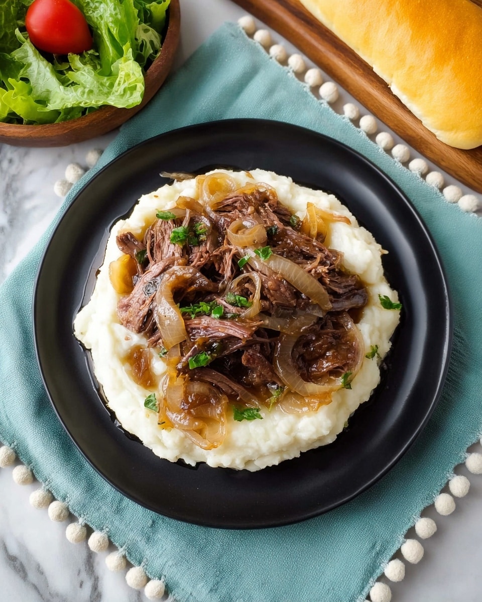 A serving of shredded beef with caramelized onions rests on a thick base of creamy mashed potatoes on a black plate. The mashed potatoes are smooth and white, spreading evenly across the bottom layer. The beef is dark brown and tender, placed on top of the potatoes with cooked, translucent onions scattered over and around it. Small pieces of chopped green herbs are sprinkled on the beef and onions for color. The plate is set on a pale blue-green cloth with small pom-pom edges, all on a white marbled surface. To the top right of the plate, a yellow loaf of bread is visible, and to the top left, a bowl of green lettuce with a single red cherry tomato is partially shown. photo taken with an iphone --ar 4:5 --v 7