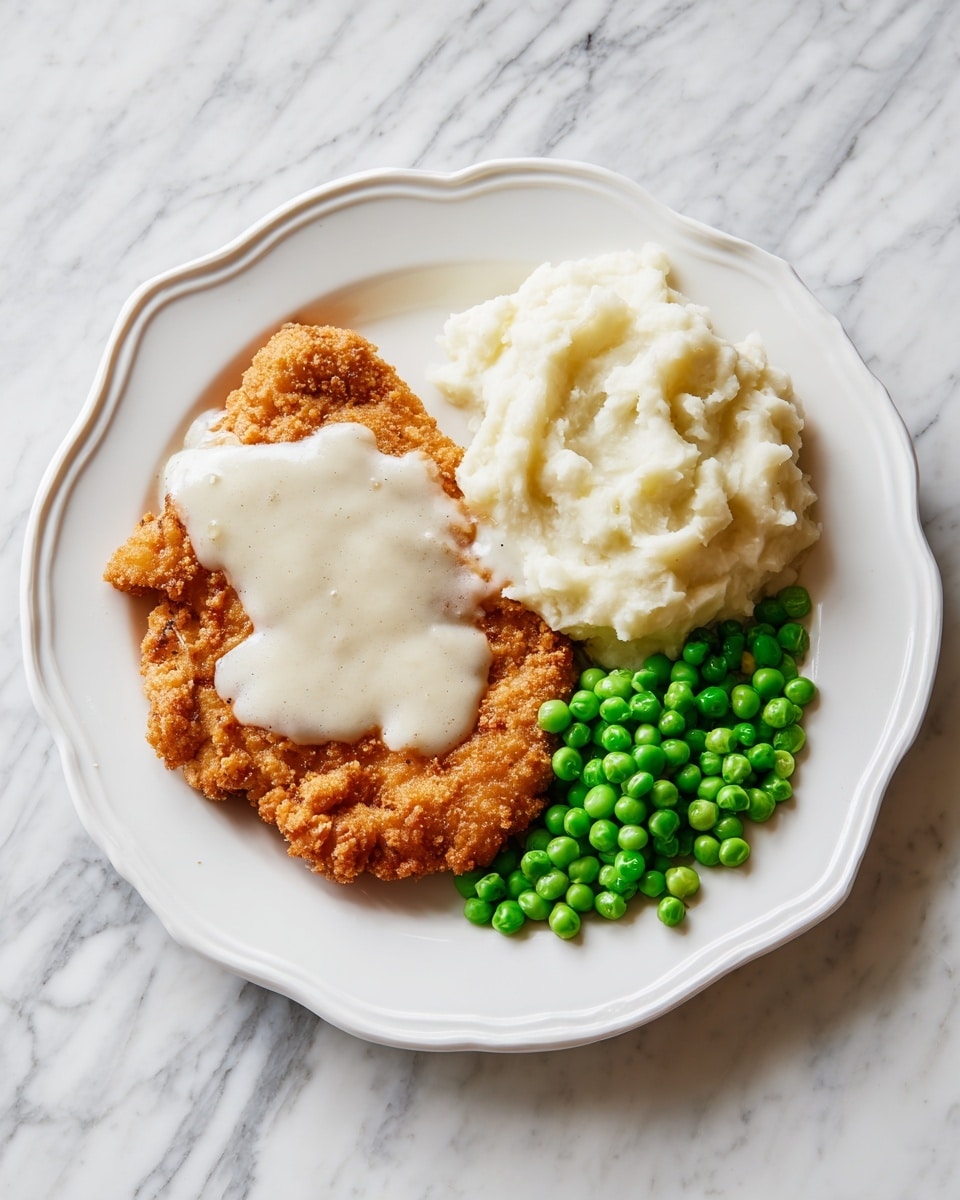 The image shows a white plate with three sections of food on a white marbled surface. The main section has a golden-brown fried chicken steak with a slightly rough and crispy texture, topped with a layer of smooth, white gravy that partially covers the chicken. To the right of the chicken is a fluffy mound of creamy, white mashed potatoes with a soft texture. Below the mashed potatoes is a cluster of bright green peas, round and fresh-looking, providing a vibrant color contrast. The plate's edge is scalloped, adding a decorative touch to the simple meal. Photo taken with an iphone --ar 4:5 --v 7