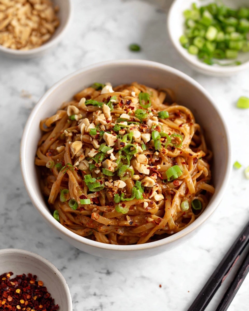 A white bowl filled with a single layer of brown noodles coated in a sauce, topped with chopped green onions, crushed peanuts, and red chili flakes scattered across the surface, sitting on a white marbled texture surface with black chopsticks nearby and small white bowls of green onions and chili flakes in the blurred background, photo taken with an iphone --ar 4:5 --v 7