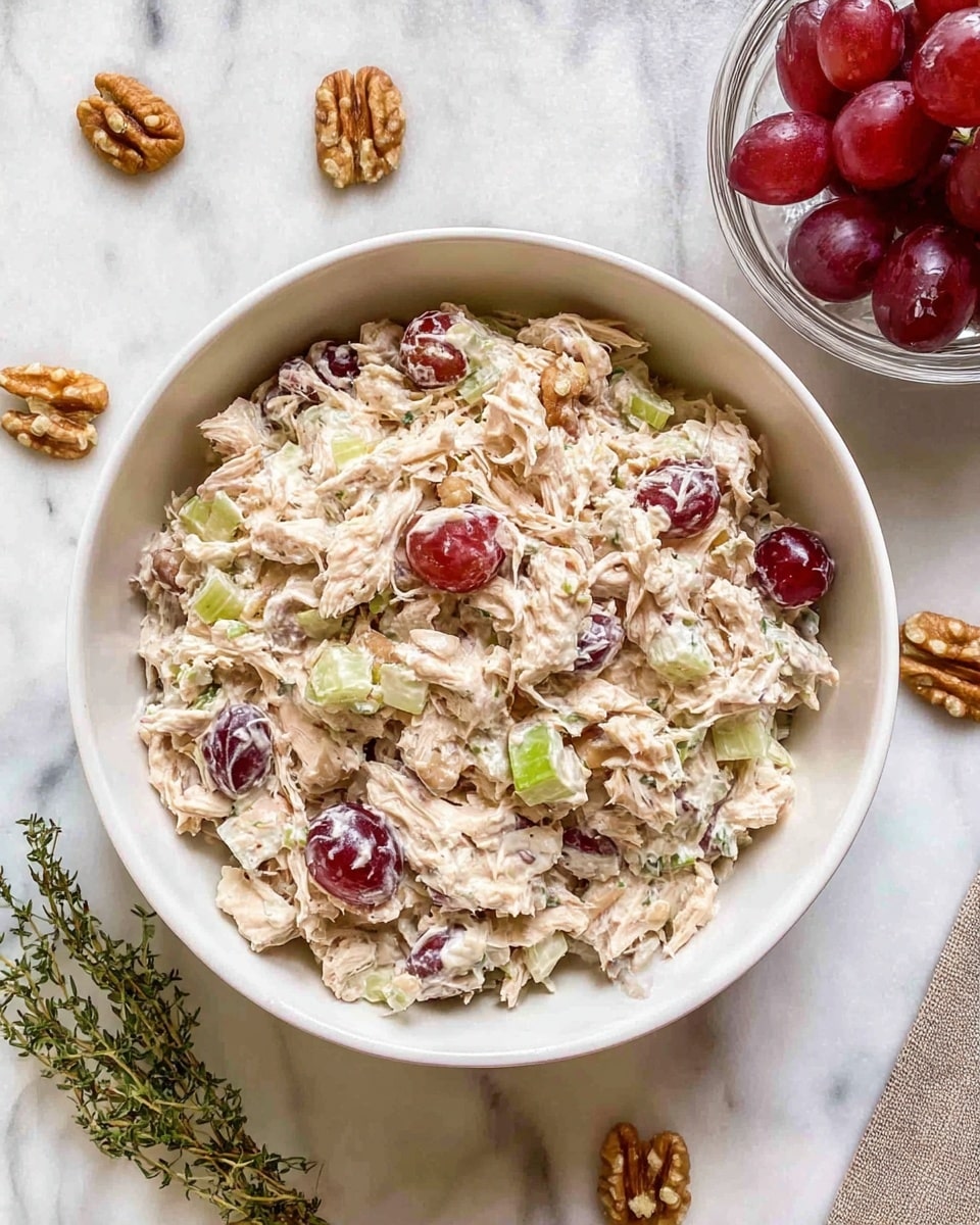 A white bowl filled with a creamy chicken salad mixed with visible red grape halves and small pieces of light green celery spread evenly throughout the dish. The salad has a thick, slightly chunky texture with shredded chicken coated in a white, creamy dressing. Around the bowl are scattered walnut pieces on a white marbled surface, with a small glass bowl of whole red grapes on the right and a sprig of green thyme on the bottom left. The photo taken with an iphone --ar 4:5 --v 7