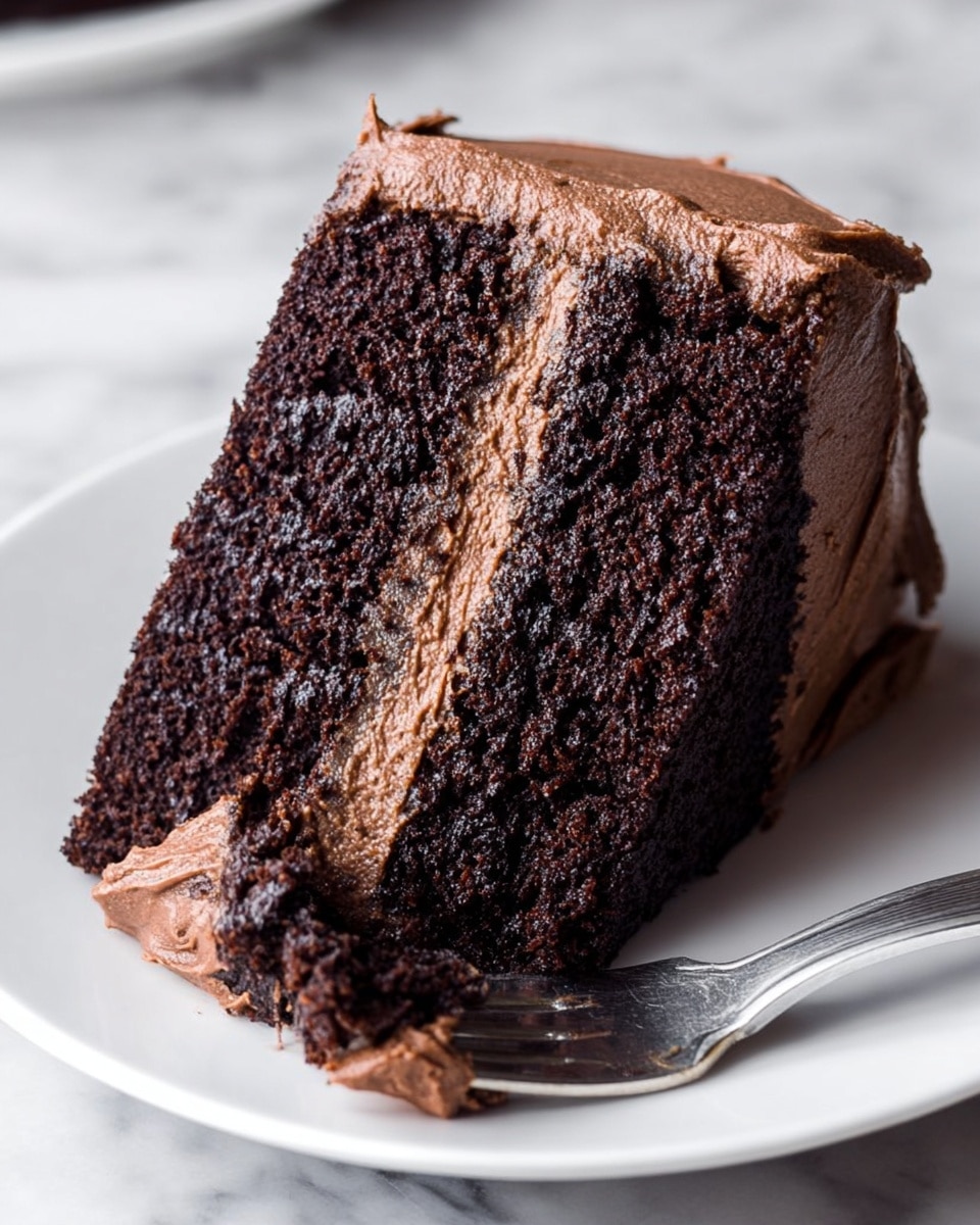 A close-up view of a two-layer chocolate cake slice on a white plate, each layer dark brown and moist with a rough texture. Between the layers and covering the outside is a thick, creamy, light brown chocolate frosting with a slightly rough but smooth appearance. A silver fork is placed on the plate, holding a small piece of cake and frosting near the bottom right side of the slice. The background is a white marbled surface. Photo taken with an iphone --ar 4:5 --v 7