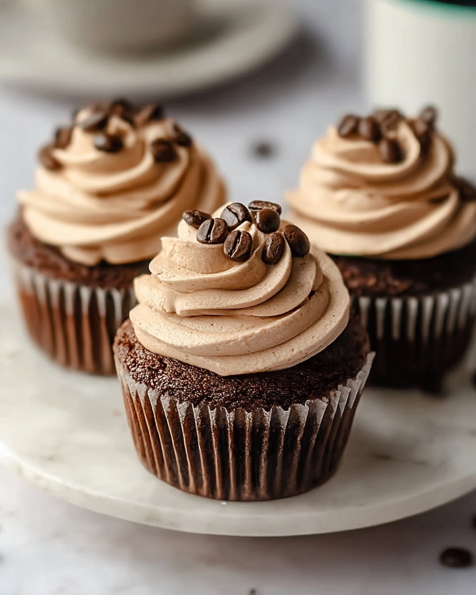 Three chocolate cupcakes sit on a white plate with a white marbled texture surface. Each cupcake has a dark, rich brown base and is topped with one swirl of light brown, creamy frosting. The frosting is thick and smooth with a slight airiness. Small, shiny dark brown coffee beans are scattered evenly on top of the frosting on each cupcake. The background is softly blurred to keep focus on the cupcakes. Photo taken with an iphone --ar 4:5 --v 7