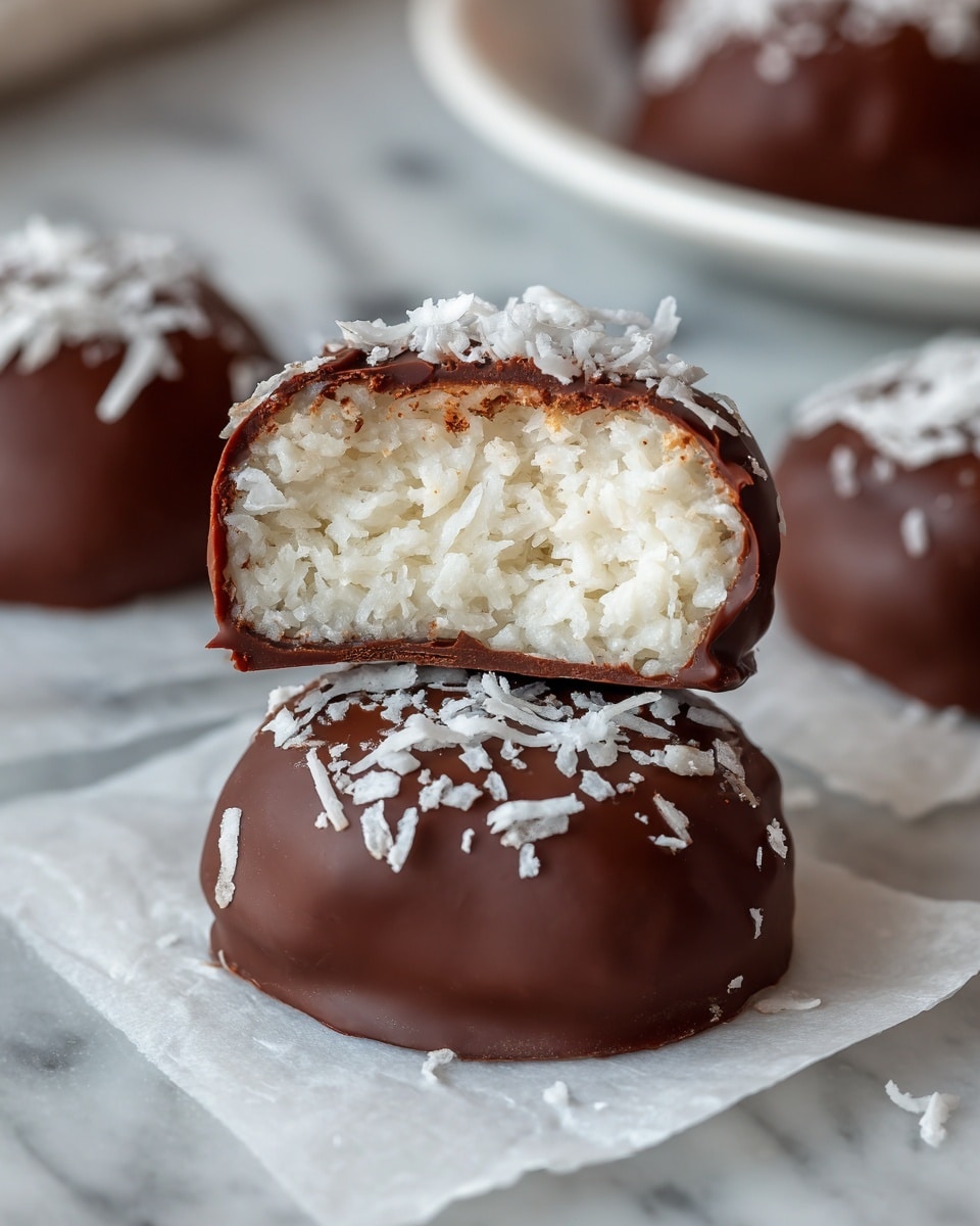 The image shows a close-up of a chocolate-covered coconut candy with one piece cut in half, revealing a thick white, shredded coconut filling inside. The chocolate coating is smooth and dark brown, forming a thin outer layer that wraps around the white coconut center. The top of the candy is sprinkled with shredded coconut flakes, adding texture and a light cream color contrast. The candy rests on white parchment paper placed on a white marbled surface, and other whole chocolate-covered pieces are blurred in the background. Photo taken with an iphone --ar 4:5 --v 7