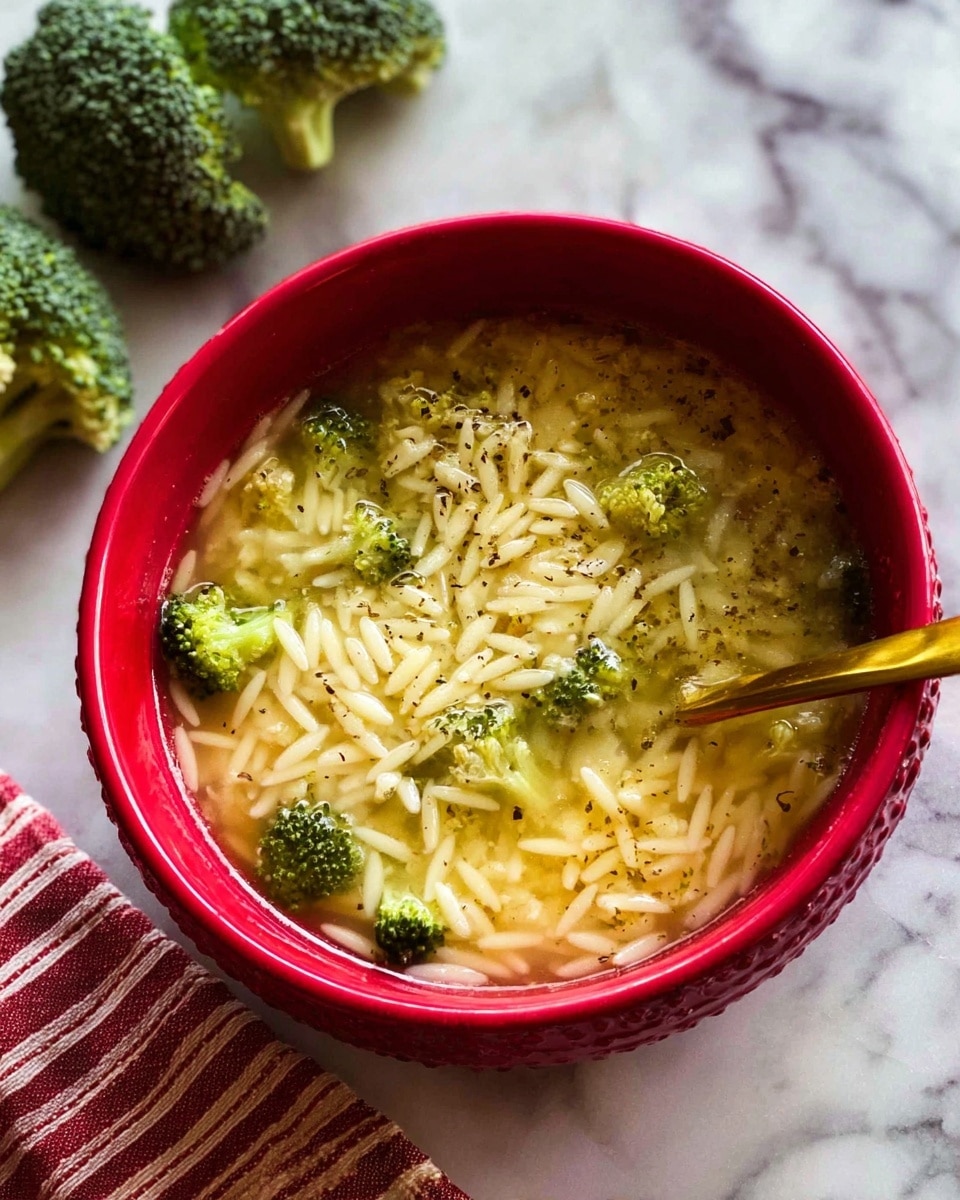 A red bowl filled with clear broth soup containing small white orzo pasta pieces and green broccoli florets floating on top; the soup is sprinkled with shredded light yellow cheese and black pepper, creating a slightly textured surface; a golden spoon is partially submerged on the right side of the bowl; in the background, there are three pieces of broccoli resting on a white marbled surface, and a red-and-white striped cloth is visible in the lower left corner; photo taken with an iphone --ar 4:5 --v 7