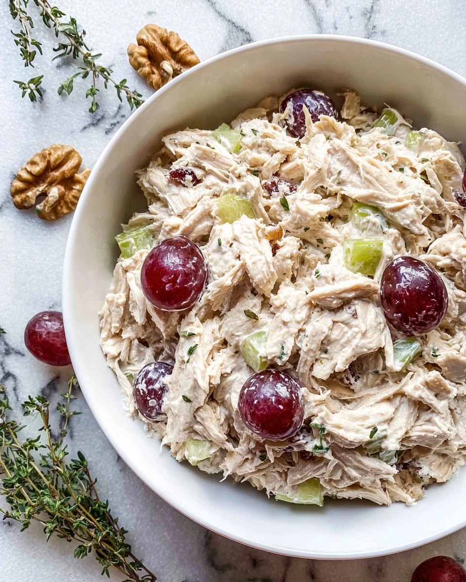 A white bowl filled with a creamy salad consisting of shredded light beige chicken mixed with sliced pale green celery and whole and halved dark red grapes, coated evenly in a thick white dressing. The salad has a chunky texture with visible layers of chicken strips and grape slices spread throughout. Around the bowl are walnut halves and fresh green thyme sprigs, all placed on a white marbled surface. photo taken with an iphone --ar 4:5 --v 7