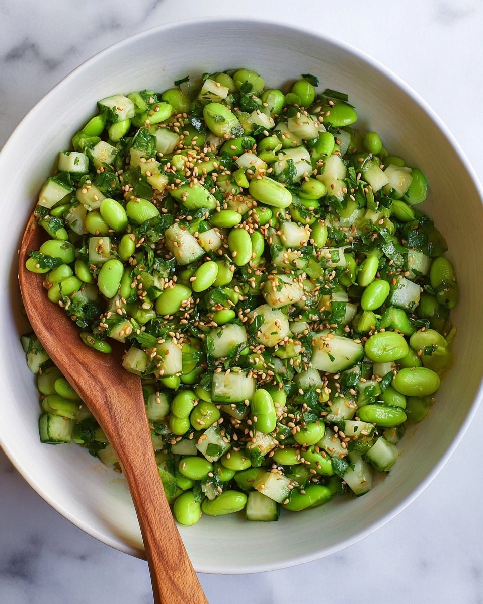 A white bowl filled with a fresh salad made of bright green edamame beans and small diced cucumber pieces, mixed with chopped green herbs and topped with light brown sesame seeds. A wooden spoon rests inside the bowl on the left side. The bowl sits on a white marbled surface. photo taken with an iphone --ar 4:5 --v 7