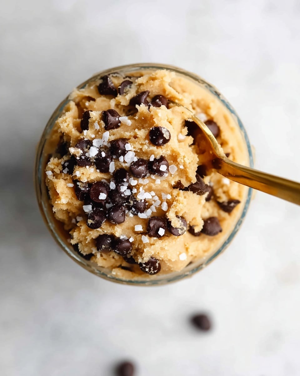 A close-up top view of a glass cup filled with three thick layers of light brown cookie dough mixed with dark brown chocolate chips scattered throughout each layer, topped with small white flakes of sea salt. A gold spoon is placed inside the cup on the right side, partially sinking into the dough. The cup rests on a smooth white marbled surface with a single chocolate chip blurred in the background near the bottom edge of the frame. Photo taken with an iphone --ar 4:5 --v 7