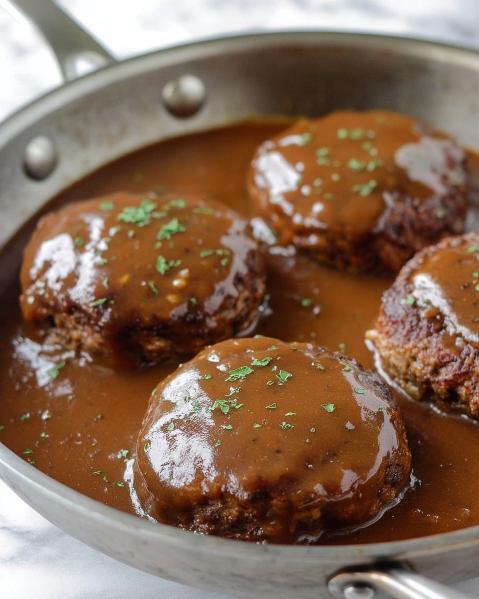 The image shows four thick, round hamburger patties covered with a shiny brown gravy, all sitting in a silver pan filled with more smooth brown gravy. The gravy has a glossy texture with small bits, and there are sprinkles of chopped green herbs on top of each patty. The patties have a slightly rough surface peeking out from under the gravy, and the pan's edge and handle are visible with a white marbled background behind. photo taken with an iphone --ar 4:5 --v 7