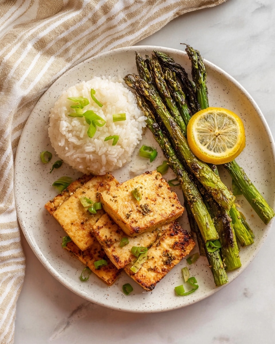 A white speckled plate holds a simple meal with three parts: at the bottom, four golden brown, pan-fried tofu pieces with a slightly crispy texture and small green onion slices sprinkled on top; on the left side, a small mound of fluffy white rice, also topped with green onion slices; on the right upper side, several charred green asparagus spears with some browning and grill marks, topped with a thin slice of yellow lemon. The plate sits on a white marbled surface with a beige and white striped cloth napkin partially visible on the upper left corner. Photo taken with an iphone --ar 4:5 --v 7