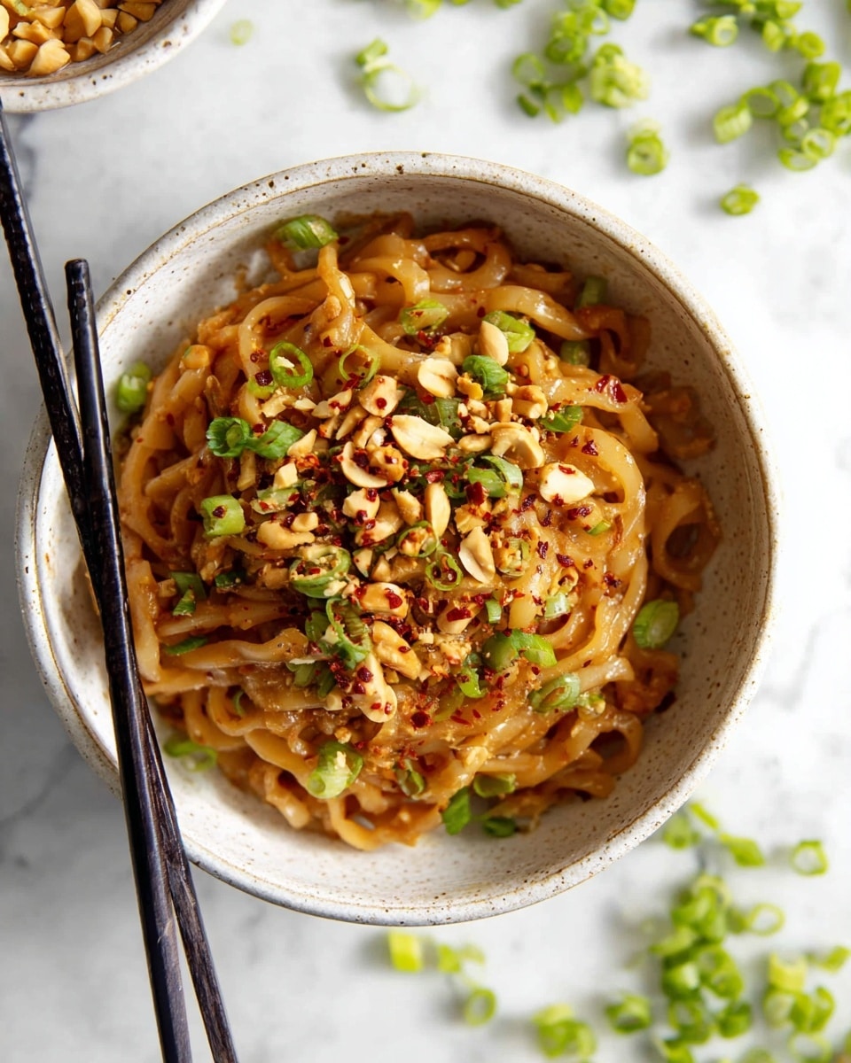 A bowl of thick, creamy brown noodles sits at the center, tangled and coated with a glossy sauce. The noodles are topped with chopped peanuts, bright green sliced scallions, and sprinkled with red chili flakes, adding pops of color and texture. Black chopsticks rest on the left side of the bowl. The bowl itself is white with a slight speckled texture, placed on a white marbled surface with scattered green onions around it. Photo taken with an iphone --ar 4:5 --v 7