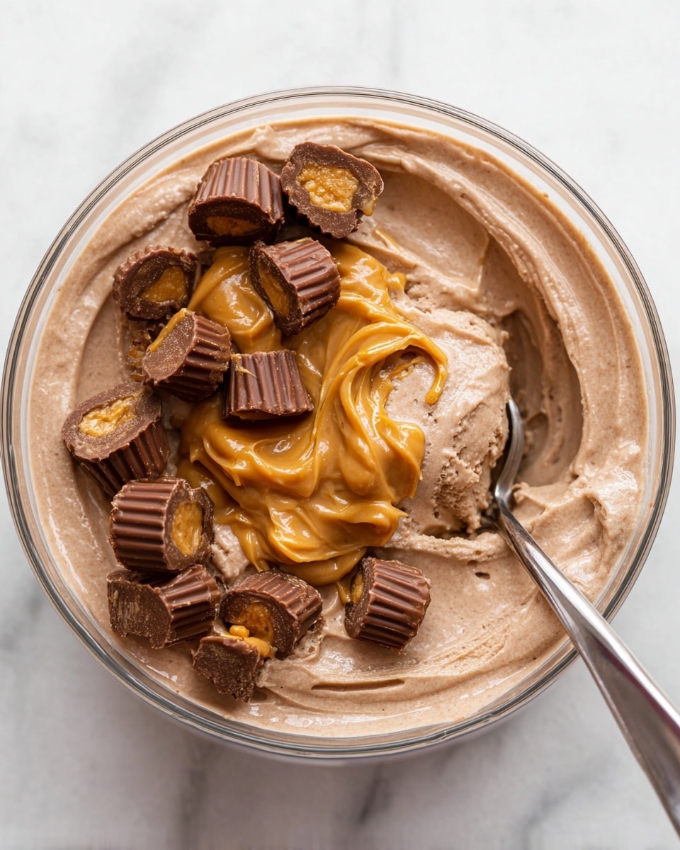 A clear bowl filled with creamy chocolate ice cream shows a smooth, light brown swirled base. On top, there is a thick dollop of shiny, golden peanut butter spread across the center. Several mini peanut butter cups, some whole and some cut in half revealing a rich peanut butter filling, are scattered over the peanut butter layer. A silver spoon rests on the right side of the bowl, partially submerged into the ice cream. The bowl is placed on a white marbled surface. photo taken with an iphone --ar 4:5 --v 7
